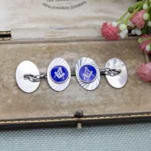 Op de afbeelding: Zilveren manchetknopen met een ovale vorm en een blauw midden met een vrijmetselaarssymbool. De manchetknopen hebben een gestructureerd, radiaal ontwerp en zijn verbonden door een ketting. Gepresenteerd in een vintage doos.