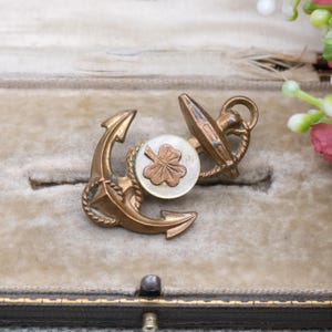 May include: Antique gold-coloured anchor brooch with a white and copper-coloured clover design. The brooch features an anchor and a circular element with a clover. The brooch is displayed in a velvet-lined box.