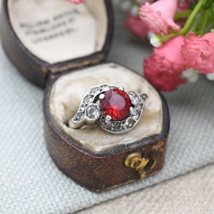 May include: A red gemstone ring with a silver band and smaller clear gemstones set around the main stone. The ring is in a small, brown velvet ring box.