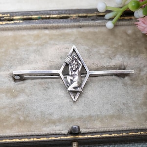 May include: A silver-toned brooch featuring a diamond-shaped design with a central figure holding a basket. The brooch has a pin closure on the back. The figure is kneeling and has a basket above its head.
