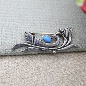 Puede incluir: Broche plateado vintage con diseño de ave estilizada. El broche presenta una piedra ovalada azul claro y detalles intrincados. Se ven las alas y las plumas de la cola del ave. El alfiler está sobre un fondo de tela neutra.