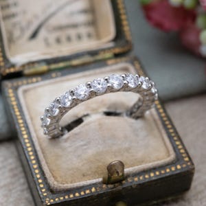 May include: A silver eternity ring with multiple round, clear gemstones. The ring is set in a vintage-style jewelry box with a light beige interior. The box is dark green with gold trim and a small clasp.