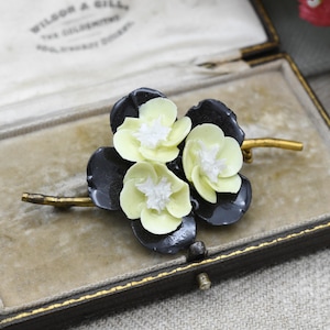 May include: A vintage floral brooch featuring three pale yellow flowers with white centers and black leaves. The brooch is mounted on a gold-toned stem and is displayed in a velvet-lined box.