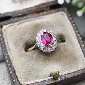 May include: A vintage ring featuring an oval ruby gemstone, encircled by a cluster of small diamonds. The ring has a gold band and is displayed in a classic jewellery box with a velvet lining.