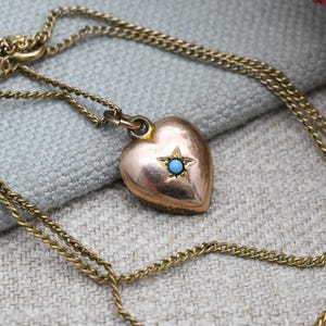 以下が含まれることがあります： 金色のハート型ペンダントネックレス。ペンダントには、中央にターコイズ色の石が入った星が付いています。ネックレスチェーンは繊細なカーブリンクスタイルです。背景は明るい灰色の生地です。