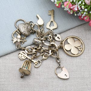 May include: A vintage charm bracelet with various gold-toned charms, including a heart, key, birdcage, and four-leaf clover. The bracelet is made of linked chains and features a mix of textures and shapes, creating a unique and detailed accessory.