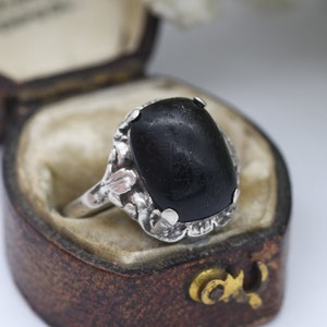 Peut inclure: Une bague en argent avec une grande pierre précieuse noire sertie dans un motif floral. La bague est dans une petite boîte brune.