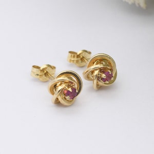 May include: Gold-coloured stud earrings with a knot design, each featuring a small, round, red gemstone. Two gold earring backs are also visible. The earrings are set against a white background.