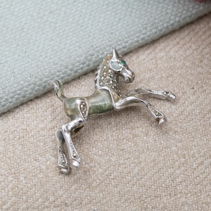 May include: A silver-toned horse brooch with a detailed design. The horse has a green-toned body and is adorned with small, sparkling crystals and a single green gem for the eye. The brooch is a decorative accessory.