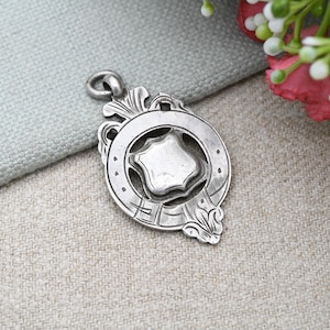 Può includere: Ciondolo argentato con un centro a forma di scudo, circondato da un anello decorativo. Il ciondolo presenta dettagli intricati, tra cui motivi floreali e geometrici. Un piccolo anello nella parte superiore per l'attacco.