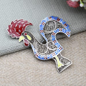 Pode incluir: Um broche de galo em filigrana de prata com esmalte azul, vermelho e amarelo. A crista e as barbelas do galo são vermelhas, o corpo é azul com flores vermelhas e as penas da cauda são amarelas.