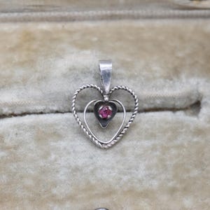 May include: A silver heart pendant with a twisted rope design around the outer heart shape. A smaller heart shape inside holds a small, round, red gemstone. The pendant has a small loop at the top for a chain.