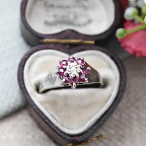 以下が含まれることがあります： 鮮やかな赤い宝石に囲まれた中央のダイヤモンドが特徴の、フローラルデザインのシルバーリング。 リングは、そのエレガントでヴィンテージなスタイルを示す、ハート型の紫色のベルベットのジュエリーボックスの中に展示されています。