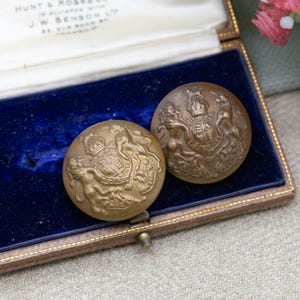 Może przedstawiać: Dwa antyczne mosiężne guziki z szczegółowymi herbami heraldycznymi, w tym lwy, korona i tarcza. Guziki są prezentowane w zabytkowym pudełku wyłożonym niebieskim aksamitem, prezentującym ich skomplikowany design i historyczne znaczenie.