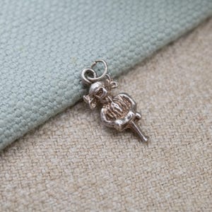 Puede incluir: Un pequeño dije plateado con forma de bailarina. El dije presenta un diseño detallado de una bailarina con tutú y un lazo en la parte superior para sujetarlo a una pulsera o collar. El dije está sobre una tela de color claro.