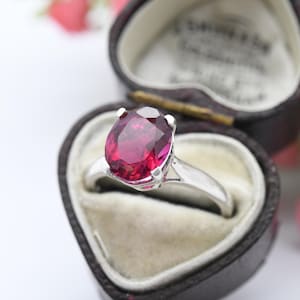 Peut inclure: Une bague en argent avec une grande pierre précieuse ovale de couleur rose foncé. La bague est dans un écrin de bague en forme de cœur.