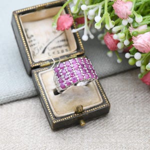 May include: A silver ring with a rectangular top, densely set with oval-cut, vibrant pink gemstones. The ring is displayed in a vintage black jewelry box with a cream-colored interior. Pink flowers and greenery are in the background.