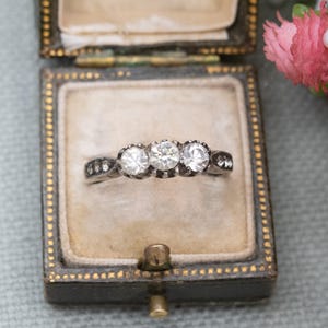 Könnte beinhalten: Ein Vintage-Silberring mit drei runden, klaren Edelsteinen. Der Ring wird in einer dunklen, verzierten Schmuckschatulle mit hellem Innenraum und goldenem Rand präsentiert. Das Design des Rings ist klassisch.