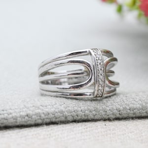 May include: A silver ring with a distinctive design. The ring has multiple parallel bands and a central section embellished with small, clear stones. The ring is displayed on a grey fabric surface, highlighting its intricate details and polished finish.