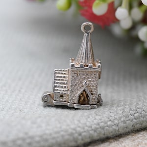 May include: A detailed silver charm in the shape of a church, featuring a steeple, arched doorway, and textured brickwork. The charm has a loop at the top for attachment and is set against a soft, neutral background.