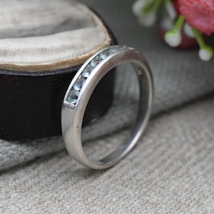 May include: A silver ring with a row of light blue gemstones. The ring is set in a channel style, with the stones flush against the metal. The ring is shown on a neutral background, with a glimpse of a jewelry box.