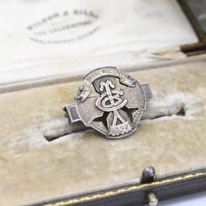 Puede incluir: Un broche plateado vintage con la inscripción "MY DEEDS WILL SPEAK" en una pancarta. El broche presenta un diseño central y el año "1937". Se muestra en un joyero forrado de terciopelo. Probablemente un artículo conmemorativo.