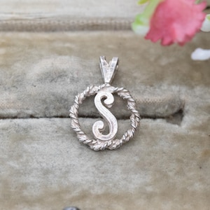 Puede incluir: Un colgante plateado con la letra "S" en cursiva, enmarcada en un diseño circular de cuerda trenzada. El colgante tiene un pequeño lazo en la parte superior para una cadena. El fondo es verde suave.