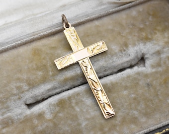 May include: Gold-coloured cross pendant with etched leaf and floral patterns. The cross has a loop at the top for a chain. The pendant is displayed against a grey velvet background, suggesting a jewellery or religious item.