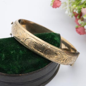 May include: A gold-coloured hinged bangle bracelet with an engraved floral design. The bracelet is resting on a dark green velvet surface. The background is white, with pink and white flowers.