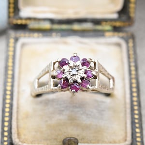 Puede incluir: Un anillo de plata con un diseño en forma de flor que presenta un grupo de pequeñas piedras preciosas púrpuras y una sola piedra blanca en el centro.