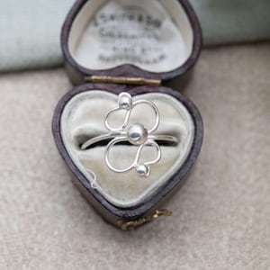 May include: A silver ring with a floral design, featuring a central sphere and curved lines with smaller spheres. The ring is displayed inside a heart-shaped, brown jewelry box with a cream-colored interior. The ring's design is delicate and modern.