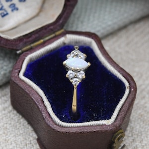 Könnte beinhalten: Ein Vintage-Goldring mit einem Opal im Marquise-Schliff, umgeben von kleinen, funkelnden Akzentsteinen. Der Ring ist in einem goldenen Band gefasst und wird in einer braunen Schmuckschatulle mit blauem Samt präsentiert. Das Design des Rings ist elegant und klassisch.