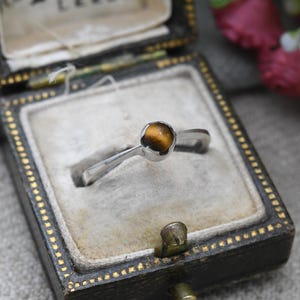May include: A silver ring with a round, amber-colored stone, presented in a vintage black jewelry box with gold trim. The ring's band has a simple, elegant design, showcasing the gemstone.