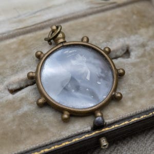May include: A vintage brass pendant with a circular frame, resembling a ship's wheel, encasing a black and white photograph. The pendant is set against a velvet-lined box, showcasing its antique charm. The photograph appears to be of a man in uniform.