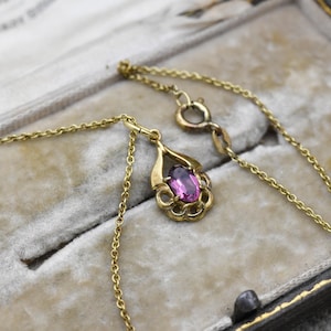 以下が含まれることがあります： 紫色の宝石が付いたペンダントが付いたゴールドカラーのネックレス。ペンダントは、装飾的なゴールドフレームにセットされた楕円形の石を持っています。ネックレスは、灰色のベルベットの裏地が付いた箱に表示されています。