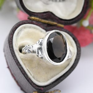May include: A silver ring with an oval, dark brown gemstone. The ring is set in a decorative silver band and is displayed in a heart-shaped jewelry box with a velvet interior.