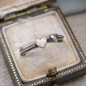 Könnte beinhalten: Silberfarbener Ring mit zwei herzförmigen Akzenten. Ein Herz ist mit einem perlmuttfarbenen weißen Material gefüllt, das andere ist massiv silberfarben. Der Ring wird in einer Vintage-Schmuckschatulle präsentiert.