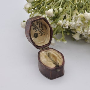 May include: A small, brown, velvet-lined ring box with a hinged lid. The inside of the lid is printed with the text "John Berry, 10211 Angel Row, Nottingham".