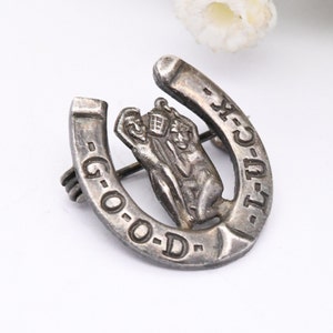 May include: Silver horseshoe brooch featuring a figure of a man holding a lantern with the inscription "Good Luck" on the horseshoe.
