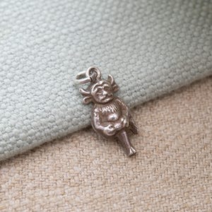 Puede incluir: Un pequeño dije plateado con forma de criatura mítica con cuernos, rostro humanoide y cola. El dije está detallado con superficies texturizadas y un lazo para sujetarlo a un collar o pulsera. El dije se muestra sobre una tela azul claro y beige.