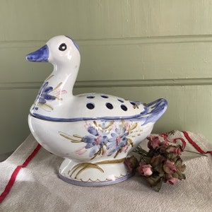 May include: A white ceramic duck-shaped flower frog with a blue beak and accents. The duck has a series of holes on its back for arranging flowers. Floral designs in blue, pink, and brown adorn the body. The duck sits on a pedestal base.