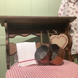 May include: A wooden shelf with a scalloped edge, featuring two copper pots hanging from hooks. A white towel is placed on the shelf, and a heart-shaped wooden sign with red accents hangs on the right side.