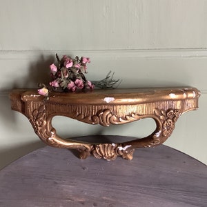 May include: A gold-painted wooden shelf with ornate detailing and a curved design. The shelf is empty, but there are dried pink flowers on the surface.