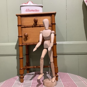 May include: A wooden artist's mannequin standing next to a small wooden nightstand with two drawers. The nightstand has a white box on top with the word "Allumettes" printed on it.