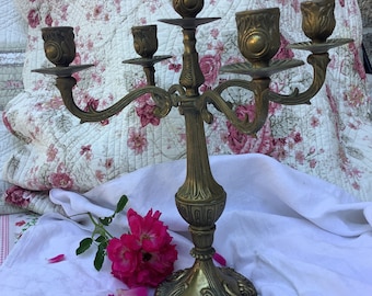 May include: A gold-toned metal candelabra with five arms and ornate details. The candelabra has a decorative base and is designed to hold candles.