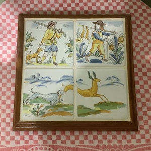 May include: A wooden framed tile set with four hand-painted tiles depicting hunting scenes. The tiles feature a hunter with a dog, a hunter with a deer, a dog chasing a rabbit, and a deer running through a field.