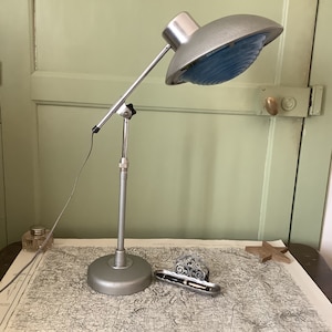 Puede incluir: Una lámpara de escritorio de plata ajustable con una pantalla azul y blanca. La lámpara está sobre una mesa con un mapa y un portaplumas de plata.