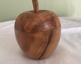 Könnte beinhalten: Eine holzfarbene, apfelförmige Box mit Deckel und Stiel. Die Box besteht aus hellbraunem Holz mit sichtbaren Maserungsmustern. Der apfelförmige Behälter steht auf einer weißen Oberfläche.