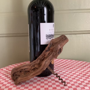 May include: A rustic wooden corkscrew with a metal worm, resting on a red and white checkered tablecloth next to a bottle of red wine.