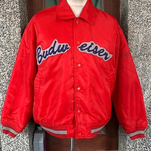Puede incluir: Chaqueta bomber de satén rojo con un logotipo "Budweiser" azul y gris en la parte delantera. La chaqueta tiene cierre de botones y puños y dobladillo de canalé.